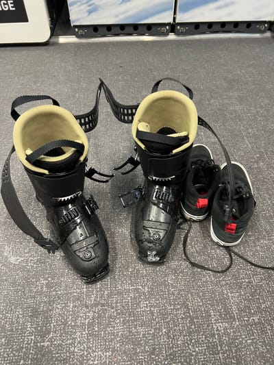 Two pairs of black ski boots with tan liners and black buckles lie on the floor of a room in Val-d'Isère, France. The room has a gray, speckled floor. The boots have straps attached. Two black sneakers are on the right side of the image, partially obscuring one of the boots. The sneakers are lace-up with red accents. The image was taken from an overhead perspective.