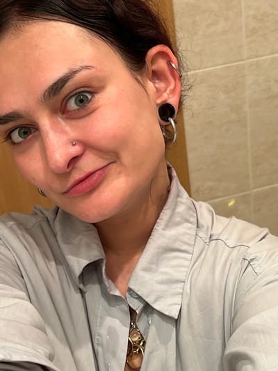 A woman is shown in a close-up, low-angle shot. Her dark hair is wet. She has fair skin, green eyes, and a nose piercing. Her ear is adorned with a large gauge earring. She wears a light grey collared shirt with a wristwatch visible near her collar. The background is a neutral beige wall. There is no indication that this image was taken in Puçol, Spain.