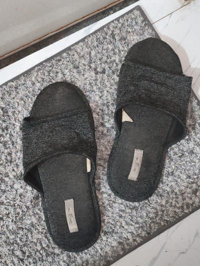 This is a top-down, eye-level, close-up shot of a pair of dark gray, textured fabric slippers resting on a gray, textured doormat. The slippers are the primary focus, with one positioned slightly to the left of the frame and the other to the right, angled slightly towards each other. Both slippers have a band across the top of the foot and a sole that appears to be made of a different material. Small, light-colored labels with text are visible on the inner side of each slipper. The doormat is situated on a white, marbled surface, likely a floor, and a portion of a metallic silver edge or frame is visible along the top left of the image. The lighting creates subtle shadows, highlighting the textures of the slippers and the doormat. The image appears to be an indoor domestic scene. There is no information in the image to suggest a location in Cavite City, Philippines.