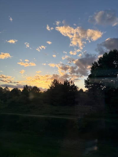 The image shows a sunset in Мажэйкаўскі сельскі Савет, Belarus. The sky is a mix of blue and orange, with scattered clouds that are lit by the setting sun. Trees are visible along the horizon line. The landscape seems to be dark and green. There are no people visible.