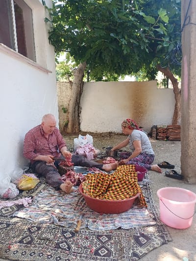 The image shows two people, an older man and a woman, sitting on a patterned rug in the courtyard of a house in Özdere, Türkiye (latitude: 38.016, longitude: 27.129). They are preparing meat, cutting it with a knife, and placing it into buckets and bowls. There are several plastic bags and a red and yellow checkered cloth on the ground. The man is wearing a red and white checkered shirt and blue jeans. The woman is wearing a white shirt and patterned pants. There is a tree in the background, along with whitewashed walls and a brick pathway. A pink plastic bucket and a red plastic basin are present. The scene appears to be in an outdoor setting.