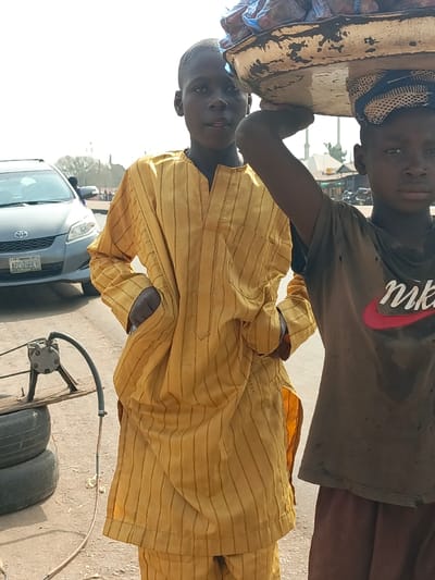 This outdoor photograph, taken in Nassarawan Eggon, Nigeria, depicts two young boys standing in what appears to be a street or roadside environment under bright daylight.
In the foreground, slightly to the left of the center, a boy in his early teens looks directly at the camera with a neutral expression. He is wearing a long-sleeved, traditional yellow garment, possibly a kaftan or dashiki, adorned with thin vertical stripes. His hands are placed in the pockets of his attire. To his right, partially obscured by the first boy, stands another boy of similar age. This second boy is engaged in street vending, balancing a large, shallow, white-painted metal tray or basket on his head, which contains items wrapped in blue plastic bags. He is dressed in a faded, dark-colored t-shirt featuring a prominent red and white "Nike" logo and looks away from the camera to the right with a serious or focused expression.
The setting reveals a dusty ground. In the left background, a silver-grey Toyota vehicle is visible, with its Nigerian license plate "ABC 299LP" clearly readable. Further to the left and lower in the frame, a makeshift piece of equipment consisting of a metallic device, possibly a small pump or engine, is set up on two stacked tires, with a hose or cable extending from it. Distant, blurred buildings and structures line the horizon, completing the scene of a bustling, semi-urban roadside. The bright, clear sky indicates midday or early afternoon.