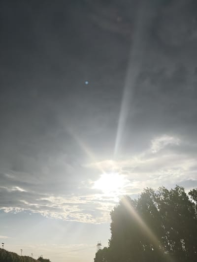 The image depicts a dramatic sky over Carinaro, Italy, dominated by the sun breaking through a dense, dark cloud cover. Sunbeams radiate downwards, illuminating patches of lighter clouds and the tops of trees silhouetted against the bright light. In the upper left portion of the sky, a small, bright blue anomaly is visible, its nature unclear. The lower portion of the image shows the tops of trees, some appearing windswept, and a faint outline of distant structures, likely part of the urban landscape of Carinaro. The overall impression is one of a dynamic and somewhat moody atmospheric condition.