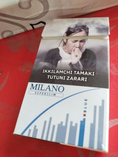 The image shows the front of a cigarette pack with a health warning. The pack is partially open and rests on a red, textured surface.
The primary subject on the pack is a black and white photograph of a young woman coughing, with smoke or vapor around her head. She is wearing a dark jacket over a lighter-colored shirt. Her expression suggests discomfort or illness.
Below the image, there is text in white, capitalized letters. The first line reads "IKKILAMCHI TAMAKI", and the second line reads "TUTUNI ZARARI". Below this, the brand name "MILANO" is prominently displayed in large, blue capital letters, with "SUPERSLIM" written in smaller, blue letters underneath.
The lower part of the pack features a stylized graphic of blue and grey vertical bars, reminiscent of a chart or graph, with a blue curved line above it. To the right of this graphic, the word "BLUE" is written vertically in blue capital letters, with a small red square preceding the word.
The setting appears to be indoors, given the lighting and the texture of the surface the pack is on. The red surface has a subtle floral pattern. The overall impression is a typical cigarette packaging design that includes a stark health warning. The text "Ikkilamchi tamaki tutuni zarari" translates to "Secondhand smoke is harmful" in Uzbek, and the location context provided is Ikkiaul, Uzbekistan.