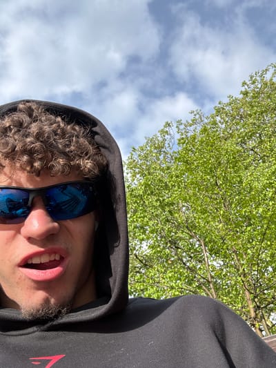 The image is a selfie taken from a low angle, looking up. A person with curly brown hair, wearing a black hooded sweatshirt and blue mirrored sunglasses, occupies the lower right quadrant. The person's mouth is open. The background shows a cloudy sky and tree branches with green leaves. Based on the reflected buildings in the glasses, I have no enough data to infer the location in the city of Aachen, Germany.