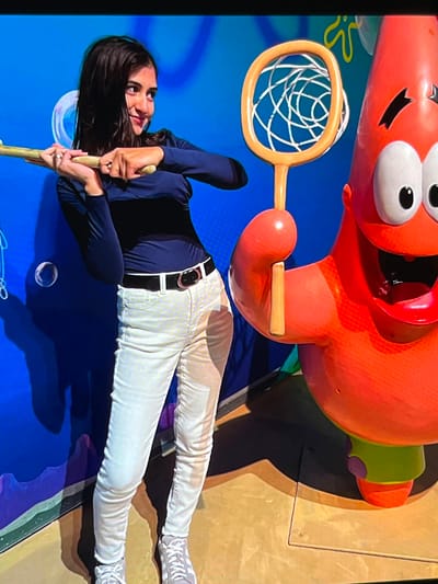 A young woman with dark hair, wearing a dark blue long-sleeved shirt, white pants, and grey sneakers, is posing for a photo. She holds a wooden stick with a net on the end, resembling a butterfly net, in front of her. To her right stands a large, inflatable or sculpted figure of Patrick Star from SpongeBob SquarePants, depicted with its characteristic orange body, large white eyes, and wide open mouth. The background is a vibrant blue wall with cartoonish bubbles and yellow starfish shapes. The floor is light brown. This scene is set in a public space, possibly an amusement park or themed attraction. The location is identified as the Netherlands, specifically the city of Nijmegen.