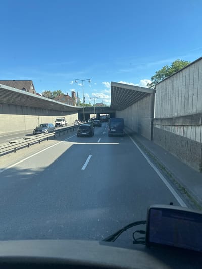 This is a photograph taken from inside a vehicle, looking forward along a multi-lane road that passes under a modern concrete structure. The road appears to be a highway or a major thoroughfare within the city of Augsburg, Germany, given the architectural style of the tunnel and surrounding buildings. The sky above is a clear, bright blue with a few wispy white clouds. Sunlight casts shadows on the road, indicating it is daytime.

Several vehicles are visible on the road, suggesting moderate traffic. On the left side, a silver Mercedes-Benz sedan is in the left lane, and further ahead, a white utility truck is in the middle lane. In the right lane, a black car is visible, and beyond it, a dark blue van. A motorcycle is also present in the middle lane. The road is paved with asphalt and marked with white lane lines. The sides of the road are lined with concrete retaining walls and some greenery.

The concrete structure overhead has an unusual, sloping design. On the left, the structure appears to cover the road and a section of adjacent land, where buildings with tiled roofs and a water tower are visible in the distance. The right side of the road features a tall, textured concrete wall. A GPS device or dashboard camera is mounted on the vehicle from which the photo is taken, its screen showing a blue display. The overall scene depicts a typical urban road environment with ongoing traffic and architectural elements characteristic of a German city.
