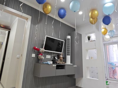 The image shows an indoor scene, likely a living room, decorated for a celebration. Several balloons, in shades of blue, gold, and white, are suspended from the ceiling. A wall with a striped grey wallpaper features a mounted flat-screen television above a grey cabinet. Two stuffed animals, a reindeer and a character with a red hat, are placed on top of the cabinet. A tall white tower fan stands to the right of the television. A doorway with a white frame is visible on the left side of the image, leading to another room. On the right, a partition wall with recessed shelving units is visible, and beyond that, a window with a view of what appears to be a balcony with laundry. The scene suggests a festive occasion, possibly a birthday party. The location is identified as Kumertau, Russia.