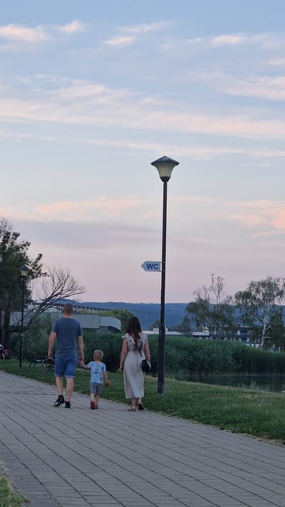 The image depicts a family of three walking along a paved pathway next to a body of water, likely Lake Balaton, in Keszthely, Hungary. The father, wearing a blue t-shirt and denim shorts, walks slightly ahead of the mother, who is in a light-colored dress. Their young son, in a patterned t-shirt and grey shorts, is walking between them, holding his father's hand. A lamppost stands to the right of the pathway, with a "WC" sign pointing to the left. The scene is set against a backdrop of lush green reeds and trees, with buildings and hills visible in the distance under a pale pink and blue sky.