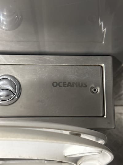 The image shows a close-up of a metal panel with the word "OCEANUS" engraved on it. To the left of the engraving is a circular, chrome-finished fixture, possibly a button or a handle. A screw with a star-shaped head is visible to the right of the engraving. The panel appears to be part of an appliance or piece of equipment, with a white ceramic-like surface visible below it, suggesting it might be a toilet or a sink area. The overall environment appears to be indoors, with artificial lighting creating reflections on the metal surface. The location in Budovo, Russia, is not evident from the image itself.