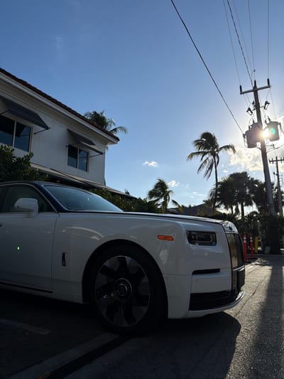 The image depicts a white Rolls-Royce Phantom luxury automobile parked on a paved surface, likely a driveway or street, in Naples, United States of America. The car is viewed from its front-left quarter, with a portion of a white two-story building featuring a red-tiled roof and multiple windows with black awnings visible to the left. Lush green foliage surrounds the base of the building. In the background to the right, a tall utility pole with numerous power lines and two transformers is silhouetted against a bright, low sun, which creates a prominent lens flare. Several palm trees are scattered throughout the scene, indicative of a tropical or subtropical environment. The sky is clear blue with sparse white clouds. Long shadows are cast across the pavement towards the viewer, suggesting either early morning or late afternoon. No people, specific actions, or interactions are visible. Two yellow and red striped bollards are partially visible behind the car on the right side of the frame. A storm drain grate is present on the pavement beneath the car's front wheel.