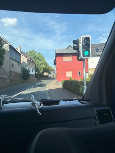 The image is a photograph taken from inside a vehicle, looking out through the windshield and side window. The view is rotated 90 degrees counterclockwise. In the foreground, the interior of the vehicle is visible on the right, with a dark dashboard and an air vent. A white cloth is tied with a rope to something in the vehicle.
The main subject of the image is a traffic light, which is illuminated with a green light. The traffic light is mounted on a metal pole. Beyond the traffic light, a street scene unfolds. A bright red building with white-framed windows and orange blinds is prominent on the left side of the street. Further down the street, more houses with traditional architecture are visible, some with stone facades and grey tiled roofs. Trees and greenery line the street, suggesting a suburban or semi-rural setting. The sky above is blue with scattered white clouds, indicating daytime and fair weather.
The activity taking place is implied by the traffic light being green, suggesting that traffic is likely moving or about to move. The location is identified as Oberwetz, Germany. There is no visible text in the image. The overall impression is a snapshot of a typical day in a German town or village, seen from the perspective of a driver or passenger.