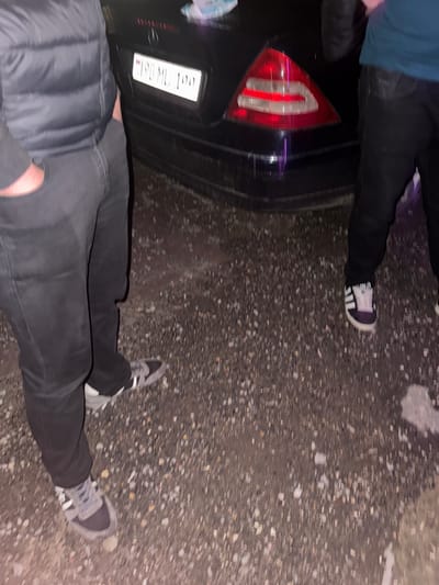 The image shows the rear of a black Mercedes-Benz car with a license plate reading "190 ML 190". Two individuals are partially visible, standing near the car on a gravelly surface, which appears to be a roadside or parking area. The person on the left is wearing a dark grey puffer vest over dark pants and grey and black sneakers. Their right hand is in their pocket. The person on the right is wearing dark pants and black and white Adidas sneakers. The car's taillights are illuminated. The scene is dimly lit, suggesting it is nighttime or dusk. The presence of a license plate format common in Armenia suggests this location is in Tumanyan, Armenia.