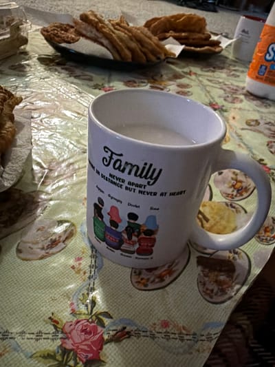 The image depicts a close-up, slightly tilted view of a table covered with a light green, patterned tablecloth. On the table are several food items, including what appear to be fried pastries or dough sticks arranged on a dark plate. A white mug is prominently featured, with a message and illustrations on its side. The mug contains a light-colored liquid. To the right of the mug, a portion of a white bottle with orange and blue lettering is visible. The background is indistinct but suggests an indoor setting. The overall impression is of a casual meal or snack setting. The specific location in Kirkland, United States of America is not discernible from the image itself.