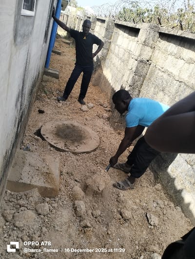 This image captures an outdoor scene in Gidan Kado, Nigeria, on 16 December 2025 at 11:29. Two adult males are observed in a trench-like area with dry, light brown, uneven soil containing scattered debris.
On the left, a light-colored concrete building wall with a visible window and a vertical blue pipe runs along its length. On the right, a weathered concrete block perimeter wall, topped with barbed or razor wire, bounds the area. Beyond this wall, trees and clear sky are visible.
An adult male, dark-skinned, wearing a black t-shirt with a small emblem and dark trousers, stands upright. He leans his right arm against the building wall, with his left hand on his hip, observing the scene to his right.
In the foreground, another adult male, dark-skinned, wearing a light blue t-shirt, dark trousers, and open-toed sandals, is crouched. He is actively engaged in breaking up the soil using a dark, cylindrical tool. His body is oriented away from the camera's direct line of sight.
Between the two main figures, a circular concrete lid, partially covered in soil, rests on the ground. The extreme foreground on the right edge of the image shows a partial view of a third individual's arm and shoulder, suggesting the perspective of the photographer.