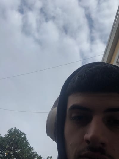 The image is oriented vertically, appearing to be captured from a low angle looking upwards. A person's face occupies the right side of the frame, partially covered by a dark hood or sweatshirt. The visible features include a dark eyebrow, an eye, part of the nose, and a section of the mouth, indicating dark hair. Below the chin, a light-colored object, possibly a cap, is partially visible.
The majority of the background is a bright, overcast sky filled with soft, diffused clouds. A thin, dark wire or cable stretches diagonally across the sky from the top left towards the bottom right. In the lower right corner, a portion of a tree with lush green foliage is visible. A small section of a light-colored building structure can be seen in the upper left corner.
The scene takes place outdoors during the daytime under cloudy weather conditions. No specific activity is clearly depicted. There is no visible text. The combination of urban elements like a building and a wire, along with natural elements like a tree and the sky, suggests an outdoor setting in Martínez, Argentina.
