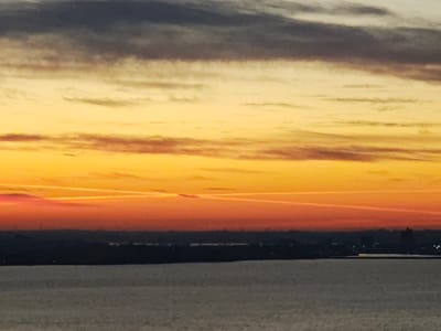 The image displays a panoramic view of a sunset or sunrise over a body of water, specifically the Great Belt, with the distant city of Korsør, Denmark, silhouetted against the horizon. The sky is characterized by distinct horizontal color bands: the uppermost section is dark grey, transitioning downwards into a broad band of pale yellow, then vibrant orange, and finally a deep crimson red along the horizon. Within the crimson and orange sky, two thin, white contrails intersect. Below the horizon, a dark, indistinct landmass, representing the cityscape of Korsør, stretches across the frame, exhibiting several sparse, faint artificial lights. The foreground is composed of a calm, greyish-blue body of water, reflecting subtle ambient light. No individuals or specific objects are identifiable within the water or landmass.