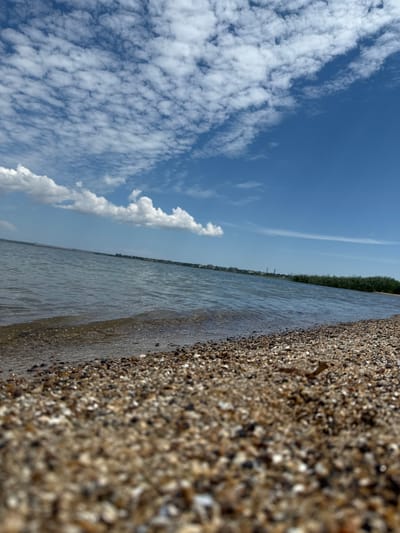 The image captures a beach scene with a body of water and a sky filled with clouds. The foreground is dominated by a textured surface of small pebbles and sand, suggesting a shoreline. To the right of the pebbles, a calm body of water stretches towards the horizon, with gentle ripples reflecting the sky. A thin line of green vegetation is visible along the water's edge in the distance, possibly trees or reeds.

The sky is a clear blue, adorned with scattered white clouds. Some clouds are wispy and spread out, while others are more defined and puffy. The lighting suggests it is daytime, likely late morning or afternoon, with the sun illuminating the scene.

The image has a tilted perspective, as if the camera was held at an angle. There is no visible text in the image. The scene conveys a sense of tranquility and natural beauty, typical of a seaside or lakeside environment. The location context of Sukholuzzhia, Ukraine, places this scene within a specific geographical area, potentially hinting at a river or coastal setting. The overall impression is one of a peaceful, natural landscape under a bright, cloudy sky.