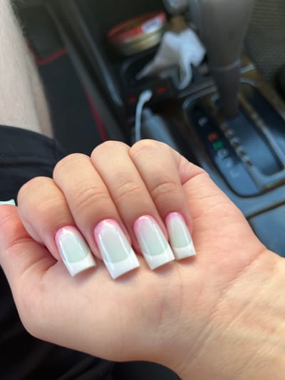 A person's hand with square-shaped, light-green gradient French tip nails is visible in the foreground of the image. The nails have a gradient effect, transitioning from a pale green at the cuticle to white tips, with a subtle pinkish hue near the tips. The background of the image shows the interior of a vehicle, specifically the gear shift console. The gear shift is in the 'P' (Park) position, indicated by the illuminated 'P' on the display. There are also visible parts of the dashboard and a hint of a person's leg and arm in black clothing on the left side of the frame. The overall setting appears to be a car interior in Konopliane, Ukraine.