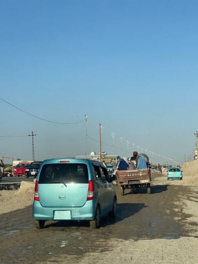 The image shows a street in Dand, Afghanistan, likely in an urban area, with a mix of vehicles and pedestrian traffic. The road surface appears to be partially unpaved and muddy. A light blue Suzuki Wagon R is the closest vehicle in the image. Further down the road, a cart is being pulled by a donkey or horse and appears to be carrying people. At least one woman is wearing a burqa. A teal-colored car is visible further up the road. In the distance, a row of light poles stretches along the road. The sky is clear and light blue.