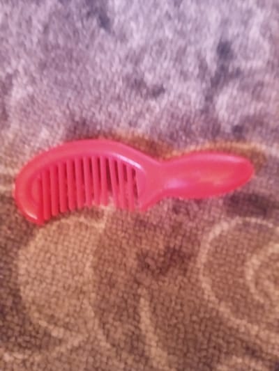 A pink comb is lying on a patterned textile surface. The comb has a curved handle and a row of teeth. The textile appears to be a rug or blanket with a textured weave. The lighting in the image is dim, and the focus is somewhat soft. There are no people or other objects visible in the frame. The image does not contain any information that would allow for the localization of the scene in Kyzyl-Sengir, Kyrgyzstan.