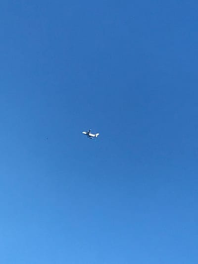 A white airplane soars against a clear, bright blue sky. Its wings are visible, along with its tail and fuselage, indicating it is in flight. A small black speck appears slightly below the plane, possibly another distant object or a speck of debris. The angle suggests the photo was taken from the ground, looking directly upward.