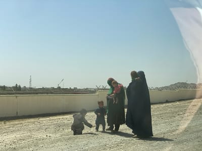 The image shows a group of five people in a rural, undeveloped area of Pir Dust, Afghanistan, located at 31.633 latitude and 65.854 longitude. There are three women wearing traditional clothing, one of whom is holding a young girl in her arms. The women are standing together, facing the camera. There are two young boys standing to their left, facing the camera. The youngest boy is crouching down with his hand outstretched. The ground is a dry, dusty dirt. A white wall with a fence, likely a security barrier, separates the group from a distant building with a communication tower. The day is clear and sunny. The image was likely taken from a vehicle, as the foreground appears out of focus and blurry.