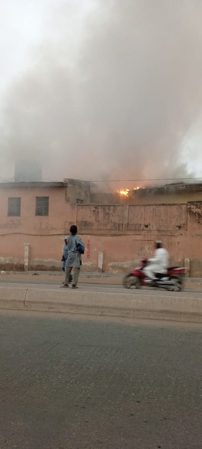 The image captures an active fire incident affecting a building, viewed from across a road. Thick, dark grey smoke fills the upper portion of the frame, billowing from a light peach-colored, possibly two-story building with a flat roof. Bright orange and yellow flames are clearly visible on the right side of the building's roofline, indicating the source of the fire. Two dark, rectangular windows are visible on the left side of the upper floor of the building. A low, concrete wall or fence runs in front of the building.
In the midground, standing on a raised curb or median separating them from the main road, are at least two individuals observing the scene. The most prominent figure is a person, likely a child or young adult, seen from behind, wearing a light blue or grey tunic and patterned trousers. They are looking towards the burning building. Another person is partially visible behind them.
To the right of the observers, on the road, a person in a light-colored, possibly white, garment and cap is riding a red motorcycle. The motorcycle and rider are blurred due to motion, suggesting they are in transit past the scene.
The foreground consists of a grey asphalt road surface. The overall lighting suggests an overcast day or possibly late afternoon, with the brightness of the flames contrasting against the smoky sky. On the wall behind the people watching, there is some faded reddish-brown vertical writing or numbering that appears to be "3+150" or similar markings.