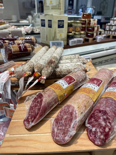 The scene depicts a detailed view of a deli counter or specialty food display within a retail establishment, likely a CONAD supermarket, located in Monsummano Terme, Italy. In the foreground, three plastic-wrapped salamis, labeled "SALAMINO TOSCANO" and displaying cut cross-sections of reddish, coarse-ground meat with white fat particles, rest on a light-colored wooden surface. Behind these, numerous whole, unwrapped dry-cured salamis with characteristic grayish-white casings and individual tags secured by red string are arranged on a wooden tiered display. Additional packaged cured meats are visible on the left side of the midground. The background features shelving units; on the right, multiple small jars of preserves or honey are stacked on miniature wooden pallets, while in the center, several tall, rectangular cardboard boxes branded "SAPORI D'TOSCANA" are stacked. A digital display showing "26.xx" is present on a lower shelf. The environment is brightly lit, consistent with an indoor commercial setting, and no individuals are visible.