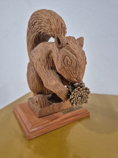 The image displays a finely carved wooden sculpture of a squirrel, depicted in a sitting posture with its bushy tail arcing over its back. The carving features textured surfaces to simulate fur, defined ears, and carved eye sockets. The squirrel clutches a small, natural pinecone between its front paws. The sculpture is mounted on a rectangular, polished wooden base with a beveled edge, which includes a small, undecipherable metallic plaque. This entire assembly rests on a smooth, reflective, golden-brown surface, likely a tabletop. The background consists of a plain, light grey wall. No visual information within the image indicates its specific location in the city of El Espinar, Spain.