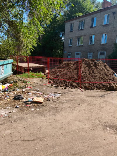 The image depicts a construction site or repair area in the city of Rubtsovsk, Russia. A large pile of dirt is cordoned off by a red metal fence. To the left of the dirt pile, a blue dumpster is visible, with a substantial amount of trash and debris scattered on the ground in front of it. In the background, a multi-story brick building with multiple windows stands under a clear sky, partially obscured by lush green trees. The ground is unpaved, appearing to be a dirt or gravel surface with visible tire tracks.