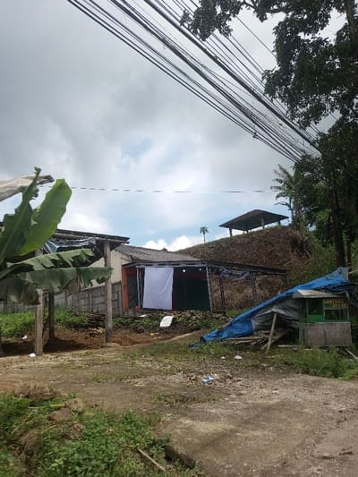 The scene is located in the city of Ura-Dozhi, Bhutan. The image depicts a daytime outdoor environment under a cloudy sky with patches of blue. In the foreground, an uneven dirt and concrete ground surface is visible, partially covered with sparse green vegetation and scattered debris. On the right, a makeshift lean-to structure, constructed from wooden poles and covered by a blue tarp and lighter fabric, stands adjacent to a small, green-painted kiosk with a white trim. This kiosk features a sign displaying green text that reads "LUNTONG SAYUR."
In the mid-ground, slightly elevated on a dirt bank, are two attached, single-story structures with corrugated metal roofs. The left structure has light-colored walls, partially obscured by large green leaves, possibly from a banana plant. The right structure, appearing to be a commercial establishment, has orange-brown walls and a vertical white fabric covering an opening. To the right of the white fabric, a dark opening is visible. A dark banner is present above this opening.
Behind these structures, a steep, barren dirt hill rises, sparsely covered with dry vegetation. On top of this hill, a small, open-sided structure with a dark, sloped roof, resembling a gazebo or shelter, is visible. A slender tree or plant stands to its left. Denser green trees line the right side of the hill. Overhead, numerous dark electrical or communication wires traverse the upper portion of the frame diagonally, partially obscured by tree branches on the upper right. No individuals are present in the scene.