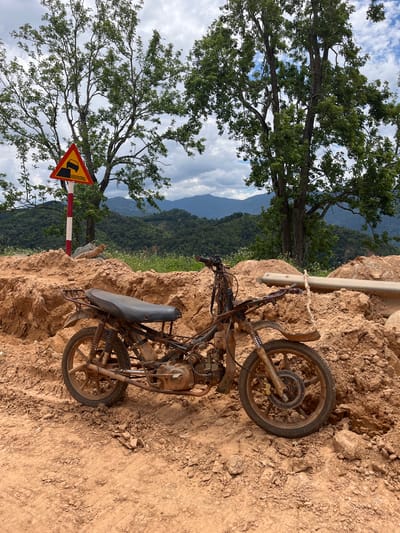 A rusty, dilapidated motorcycle sits abandoned on a dirt pile in a rural, mountainous landscape. The motorcycle is incomplete, with its front fairing, handlebars, and side panels missing. Its tires are worn, and the frame is coated in a thick layer of reddish-brown dirt. The surrounding environment is characterized by a steep, uneven terrain with large mounds of earth, possibly from construction or excavation. In the background, lush green hills roll into the distance under a cloudy sky. A yellow triangular road sign with a black symbol depicting a sharp descent or drop-off is visible to the left of the motorcycle, mounted on a striped red and white pole. The scene suggests a remote location in Nao Sẻ, Vietnam, likely a construction site or an area undergoing development, where the motorcycle has been left behind due to its condition or the challenging terrain. No people are visible in the image.