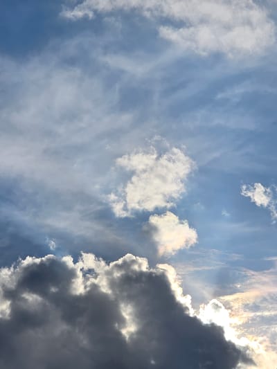 A low-angle, vertical shot captures a dynamic sky over Kostelec na Hané, Czechia. The scene is dominated by a dramatic interplay of light and shadow amongst the clouds. On the right side of the frame, thick, dark storm clouds are edged with brilliant sunlight, suggesting the sun is either rising or setting. Beams of light pierce through the darker clouds, creating a dramatic, almost theatrical effect.

Scattered across the blue sky are lighter, wispy clouds that contrast with the heavier storm clouds. Some of these clouds are illuminated by the sun, taking on a soft, golden hue, while others appear white and fluffy. The overall impression is one of changing weather, possibly an approaching or receding storm, illuminated by a powerful sun. The specific time of day is not explicitly stated but implied by the strong backlight and dramatic cloud formations. There is no visible text in the image. The setting is an open sky, devoid of any terrestrial elements, focusing entirely on the atmospheric conditions.