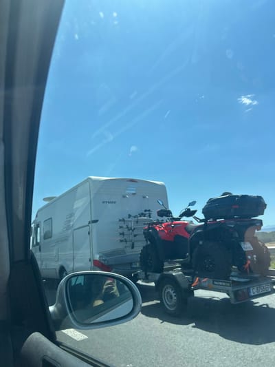 The image depicts a view from inside a vehicle, looking outwards at a sunny sky and a road. In the foreground, a silver side-view mirror reflects the face of the person taking the photo, who is holding a phone. To the right of the mirror, a trailer is visible, carrying two red ATVs. The ATVs are equipped with black storage boxes on the rear. Behind the trailer, a white recreational vehicle (RV) with the brand name "HYMER" is parked on the right side of the road. The RV appears to have a satellite dish mounted on its roof and a bicycle rack on the back. Further down the road, other vehicles are present. The environment suggests a rural or suburban setting, possibly a highway, given the presence of multiple vehicles and the open sky. The license plate on the trailer reads "C 8758 EP". The license plate on one of the ATVs is partially visible and appears to be "PK 04250". The scene is consistent with a road trip or travel scenario. The location is stated to be the city of Vakarel, Bulgaria.