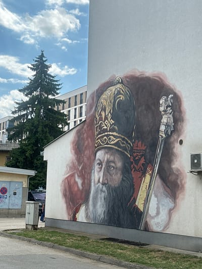 A mural of a bearded man wearing a golden, jeweled headdress and a red and gold robe is painted on the side of a white building. The man holds a silver staff in his right hand. The mural is located on the exterior wall of a building in Doboj, Bosnia and Herzegovina, near a street with a curb and a patch of grass. In the background, a tall green coniferous tree stands in front of a modern white building with many windows. A grey electrical box is positioned on the ground in front of the mural. An air conditioning unit is mounted on the wall to the right of the mural. The sky is blue with white, scattered clouds.