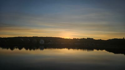 The image depicts a serene waterfront view of Södertälje, Sweden, during a sunset or sunrise. The sky is a gradient of deep blue at the top, transitioning through shades of orange and yellow due to the setting or rising sun. Wispy clouds are visible, catching the light.

In the middle ground, a silhouette of the Södertälje coastline is visible. There are several buildings, some appearing to be multi-story residences or businesses, nestled along the elevated terrain. A prominent white building with multiple windows stands out amongst darker structures. The treeline forms a dark, undulating silhouette against the horizon.

The foreground is dominated by a calm body of water, likely Lake Mälaren or the Södertälje Canal, which reflects the colors of the sky and the shoreline structures. The reflections are soft and slightly distorted, adding to the tranquil atmosphere. The overall impression is one of peace and natural beauty. There are no people or animals clearly visible in the image.