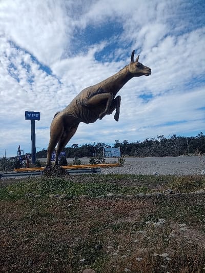 A kangaroo statue stands against a cloudy sky. The statue is a golden-brown color and appears to be mid-leap. A YPF gas station sign is near the kangaroo. The kangaroo and the sign sit on a patch of grass in front of a gravel area and a line of trees.