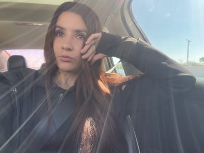 A single female subject is depicted seated within a vehicle's interior. She has long, dark brown hair with reddish undertones and is wearing a black zippered jacket over a dark top, secured by a black seatbelt across her chest. Her left hand, adorned with a ring on the fourth finger, is positioned with fingers touching her temple. Her head is slightly tilted, and her eyes are directed upwards and to the right. Strong sunlight enters the vehicle from the right, causing prominent lens flare across the frame. Through the passenger-side window, a clear blue sky, a light pole, and distant foliage are visible, indicating an outdoor environment in Fraccionamiento Los Heroes Queretaro, Mexico.