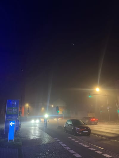 The image depicts a heavily foggy night scene on a street in Bamberg, Germany. Dense fog obscures visibility, causing all artificial light sources, including streetlights, traffic lights, and vehicle headlights/taillights, to create prominent halos and lens flares.
On the right side of the frame, a dark-colored car is visible, its taillights glowing in the fog. The license plate, "BAM TK 116", confirms the location in Bamberg. Another vehicle with red taillights can be seen further down the road, partially obscured by the haze. Multiple traffic lights are visible throughout the scene, mostly displaying green, indicating an active intersection.
Along the right edge, a brick-patterned sidewalk runs parallel to the road. A prominent blue and white illuminated sign stands beside the sidewalk, displaying brand names: "ARAL", "REWE TO GO", and "Ultimate", along with a downward-pointing arrow and the word "SuperCard". Faint outlines of buildings, including one with the partially visible text "ANNABERG", are discernible through the thick fog in the background. The time is clearly night, and the primary notable detail is the extremely foggy weather conditions. No individuals are clearly visible.