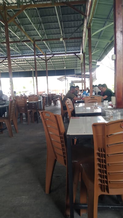 The image depicts an outdoor seating area of a restaurant or food court in Keude Trienggadeng, Indonesia. The scene is covered by a metal roof supported by wooden and metal beams, with some natural light filtering in. Numerous tables and brown plastic chairs are arranged throughout the space. In the background, several people are seated at tables, some eating or conversing. The flooring appears to be concrete. The overall impression is of a casual dining establishment.