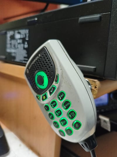 A close-up, angled shot shows a white Motorola radio microphone with green illuminated buttons. The microphone is held in front of a dark electronic device, possibly a base station or computer, with some of its screen visible. The background is out of focus, with a wooden surface in the foreground and hints of other office equipment. The microphone appears to be used, with some dust and grime on its surface. The scene suggests a professional or industrial setting, likely in an office or control room environment. The location in Tukums, Latvia, is not discernible from the image itself.