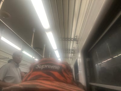 The photo shows an interior space, possibly a train or bus. Rows of fluorescent lights run along the ceiling, illuminating a grey floor with circular indentations. Overhead storage racks are visible, along with a handrail supported by slender metal poles. Two people appear in the shot. One wears an orange and black zebra-striped Supreme shirt and another is wearing a white shirt. The image is rotated.