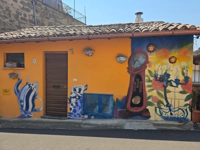 This is a medium shot, eye-level, exterior photograph of a building with vibrant, surrealist murals painted on its orange and blue-painted facade. The building has a terracotta tiled roof and a wooden door. The mural depicts a stylized grandfather clock, teacups and a teapot, and a floral pattern with sun decorations. A cactus in a terracotta pot is visible to the right. The scene is set on a paved street under a clear blue sky. This mural is located on Via Cardinal Verri in Viterbo, Italy.
