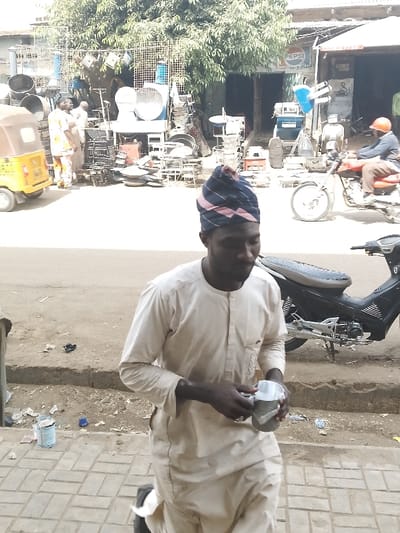 The scene depicts a street in Kofar Mazugal, Nigeria. In the foreground, a dark-skinned male, wearing a light-colored traditional tunic and a blue and pink striped cap, walks towards the left on a paved sidewalk. He holds a small cup or container, enclosed within clear plastic, in his hands. A black motorcycle is parked on the sidewalk to his right. A partially open paint can and scattered debris are on the ground near the sidewalk curb. Across a dusty road, a yellow Keke Napep is partially visible on the far left. Two males, one in a patterned tunic, stand near an open-front shop on the opposite side of the road, which displays numerous metal kitchenware items such as large pots, basins, and industrial fans. A large green tree provides shade over this shop. To the right, an adjacent building features a "PEPSI" sign. A person wearing an orange helmet rides a red motorcycle away from the camera on the road. The environment suggests a commercial district.