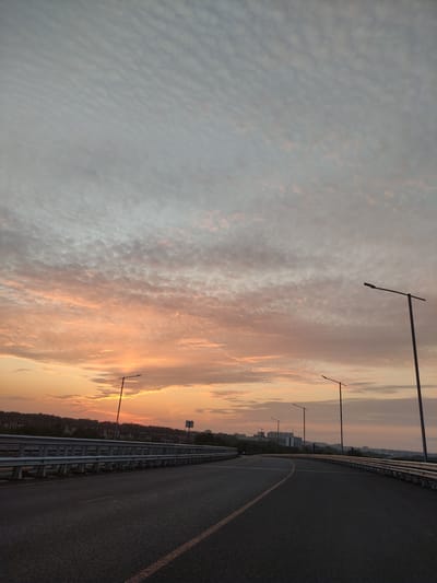 The image depicts a perspective view of an elevated asphalt road, likely a highway or bridge, under a sky filled with a gradient of colors from soft orange and pink near the horizon to a pale grey towards the top. The sky features numerous small, patchy clouds that create a textured appearance. Along the right side of the road, a metal guardrail runs parallel, and several tall streetlights with a minimalist design are visible. On the left side of the road, a similar guardrail is present, with a line of trees and buildings forming a distant landscape. The road itself is dark asphalt with a single, thin orange line marking the lane. The lighting suggests either dawn or dusk. The scene appears to be in a semi-urban or suburban environment. Given the context of Kraskovo, Russia, this is likely a road passing through or on the outskirts of the town, potentially near residential areas or undeveloped land adjacent to the town. No people or specific vehicles, other than a faint outline of a car in the distance on the left, are clearly identifiable. The overall impression is one of a quiet, transitional time of day on a modern road infrastructure.