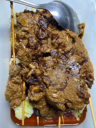 The image presents a close-up, top-down view of a traditional Indonesian dish, likely a type of satay such as Sate Matang, served in a clear rectangular plastic container. The main subjects are multiple skewers of meat, heavily coated in a thick, rich, dark brown sauce. The sauce appears to be savory and complex, with visible specks of red (possibly chili), and chunks that resemble fried shallots or caramelized onions are mixed throughout and garnish the top.

The skewers themselves are made from light-colored wood or bamboo. Beneath the meat skewers, pieces of white, compressed rice cakes, commonly known as lontong or ketupat, are visible. These rice cakes are partially submerged in a reddish-brown liquid pooled at the bottom of the container, which might be oil from the sauce or an additional chili-based condiment. A metal spoon or ladle rests on the left side of the container, partly covered by the sauce, with the word "STAINLESS" faintly visible on its handle.

The scene captures a prepared meal, suggesting it's ready for consumption or packaged for takeaway. The focus is entirely on the food, highlighting its texture and rich appearance. The context of Lampeudaya, Indonesia, aligns with the visual characteristics of this type of regional satay. No specific activity is taking place, nor are time of day, weather, or emotions discernible from this still-life food photograph.