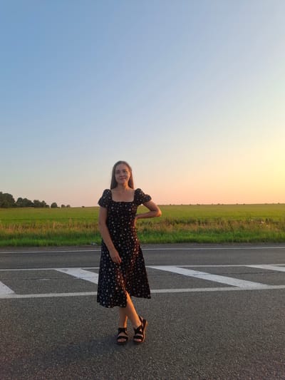 A young woman stands on an asphalt road with white markings, facing the camera. She is wearing a black floral-patterned dress with short sleeves and sandals. Behind her, a green field stretches to the horizon, with trees lining the far edge. The sky above is clear and transitions from light blue to a soft orange and yellow at the horizon, indicating sunset. In the distance, hay bales are visible in the field. The scene appears to be located in a rural or semi-rural area, possibly on the outskirts of Perevoznoe, Russia, given the vast open fields and clear skies.