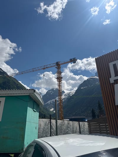 The image depicts a construction scene in Dombay, Russia, under a bright blue sky with scattered white clouds and sun glare. In the foreground, the white roof of a car is visible. To the left, a teal-colored temporary building is partially shown. A tall yellow construction crane dominates the middle ground, extending across the frame. In the background, majestic snow-capped mountains rise, covered in green forests on their lower slopes. A wooden fence and a large wooden sign with Cyrillic lettering are positioned on the right side of the image. The scene suggests ongoing development in a mountainous, possibly resort, area.