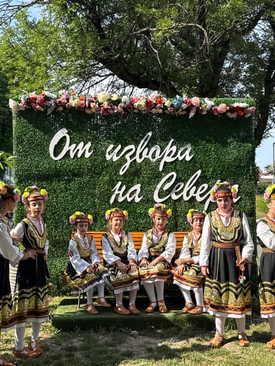 In Pordim, Bulgaria, on a sunny day, several young girls in traditional Bulgarian folk costumes are posed in front of a large green hedge wall adorned with a colorful floral arrangement at the top. The wall has white, cursive lettering that reads "От извора на Севера" (From the Source of the North). Some girls are seated on a wooden bench, while others stand. They wear embroidered vests over white blouses, with dark skirts or dresses featuring intricate gold and black patterns. All the girls have floral headbands. The ground is covered in grass, and trees provide a shaded backdrop.